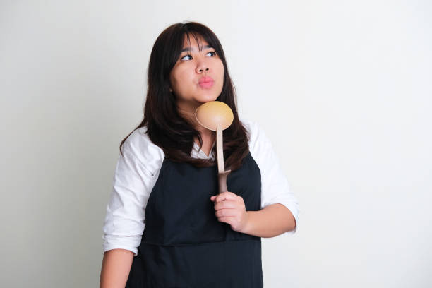 Adult Asian women wearing apron showing thinking expression while holding ladle Adult Asian women wearing apron showing thinking expression while holding ladle serving utensil stock pictures, royalty-free photos & images