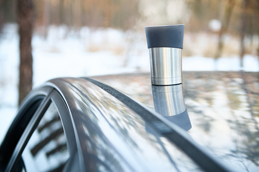 Close-up of a stainless steel thermal mug on the roof of a car in a snow covered woodland