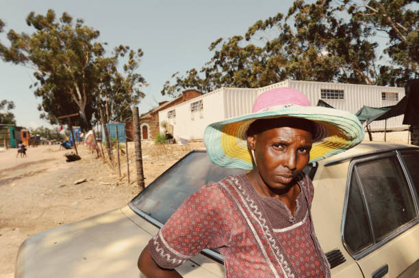 Klipheuwel, Cape Town, Western Cape, South Africa. 13 January 2016. A resident waits next to an old BMW wearing a hand-made hat.With the upcoming municipal elections approaching, many people living in under-serviced communities like  Klipheuwel informal settlement are the forgotten people of greater Cape Town.