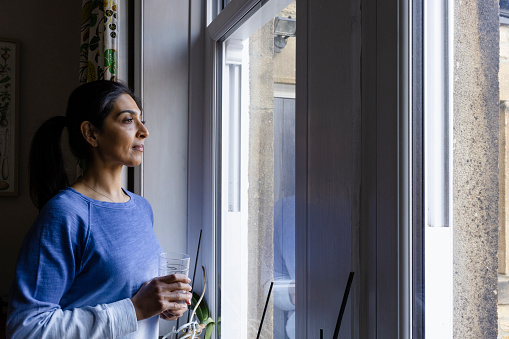 Woman standing in her home looking out of her double glazed window with a contemplative expression. She is in the Northeast of England and is holding a glass of water.