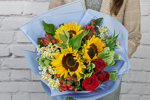 a bouquet of yellow sunflower, white daisies and red roses in a delicate blue package in the hands of a girl without a face on a light background