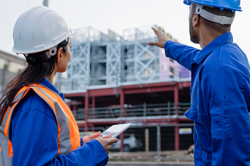 Team of mixed age and ethnic construction/engineer workers busy in the North East of England. They are wearing hard hats and overalls, talking between themselves in front of a building site. The are using a digital tablet and are pointing/gesturing to something.