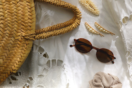 White linen lace shirt, round straw bag, sunglasses and cute hair accessories on wooden background. Flat lay.