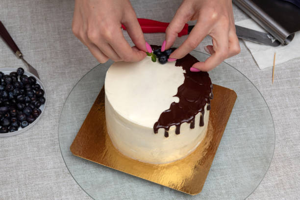 istock Women's hands decorate white sponge cake with chocolate streaks on top with mint, blueberries. Image for website about food, confectionery. Selective focus. 1388227669