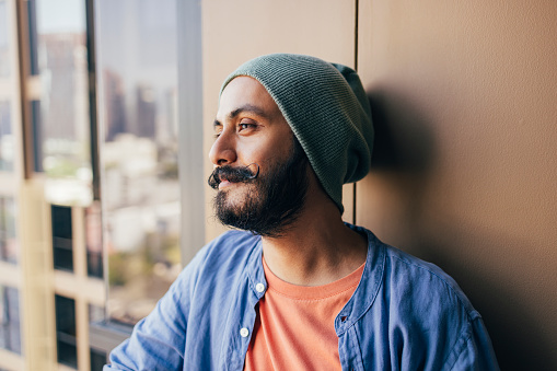 A handsome Indian male hipster with a green beanie looking away while resting on the balcony of his apartment.