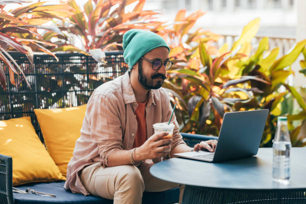 A Happy Casually Dressed Man With Moustaches Drinking Coffee While Sitting Outdoors In A Comfortable Chair And Working On His Computer A handsome Indian male businessman with glasses and a green beanie relaxing while scrolling down on his laptop. digital nomad stock pictures, royalty-free photos & images