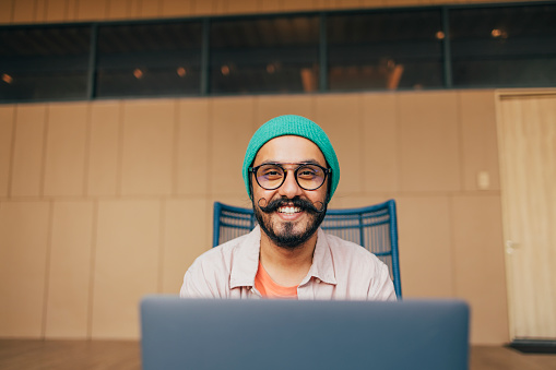 A handsome Indian male freelancer with glasses and a green beanie sitting in his new office and working on his laptop.