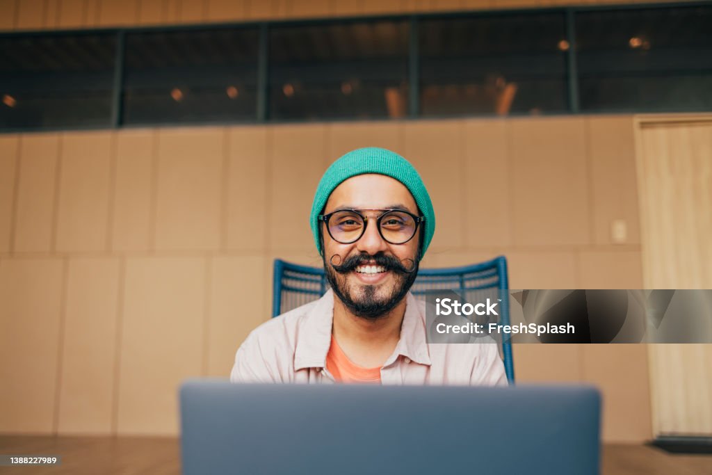 A Portrait Of A Happy Casually Dressed Man With Moustaches Looking At Camera While Sitting In His Office At Computer A handsome Indian male freelancer with glasses and a green beanie sitting in his new office and working on his laptop. Computer Programmer Stock Photo