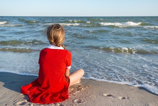 Teenager Girl in a red dress sitting on the beach dreaming and looking at the sea. Summer Vacation or holiday concept