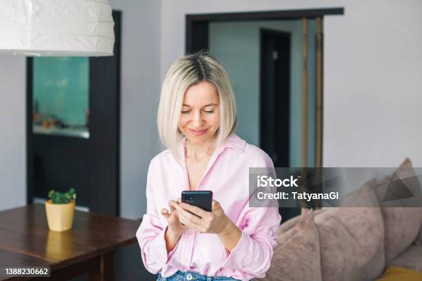 Attractive Middleaged 40s Woman Looking At Her Smart Phone And Smiling While Standing At Home Stock Photo - Download Image Now
