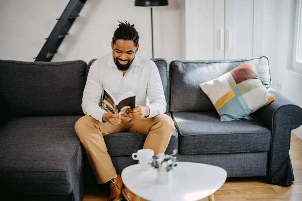 istock Positive black man reading book while sitting on sofa at home. 1388235333