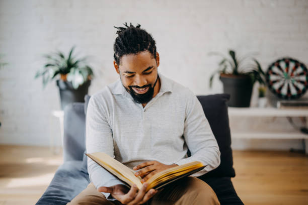 istock Positive black man reading book while sitting on sofa at home. 1388235349