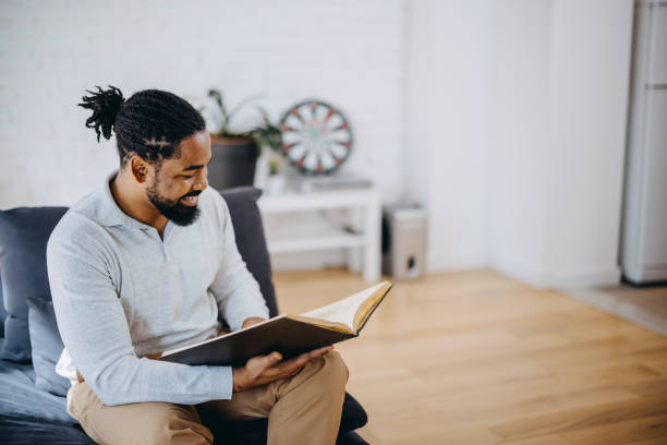istock Positive black man reading book while sitting on sofa at home. 1388235352