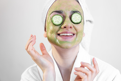 Young woman with green face mask and fresh cucumbers in white bathrobe, beauty and skin care concept.