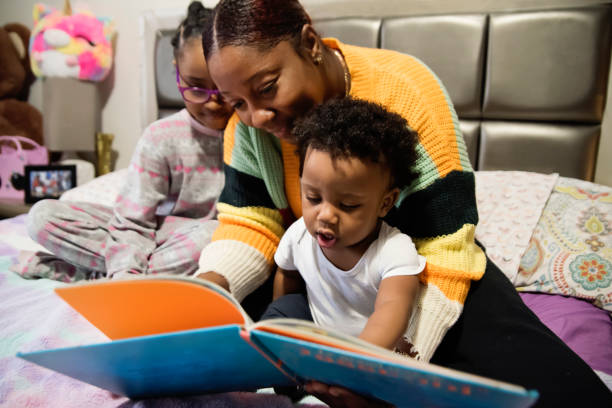 madre leyendo un libro a niños pequeños en una cama en casa. - 6 11 meses fotografías e imágenes de stock