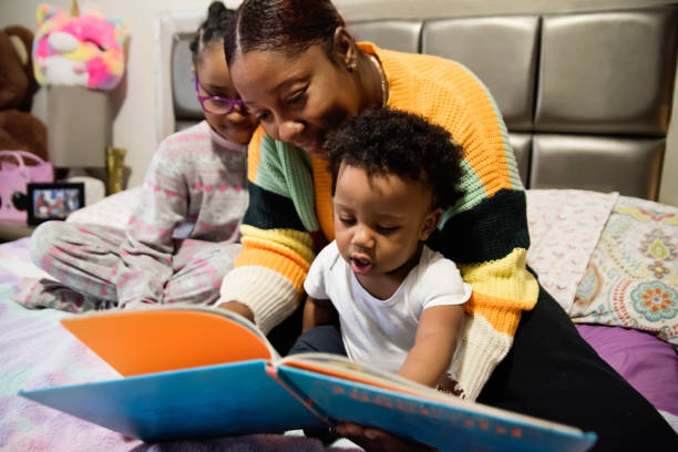 Mother reading a book to small children on a bed at home. Mother reading a book to small children on a bed at home. Mom is in her thirties, daughter is 7 years old and baby boy is 11 months. Young daughter wears eyeglasses. Horizontal indoors waist up shot. 6 11 months stock pictures, royalty-free photos & images