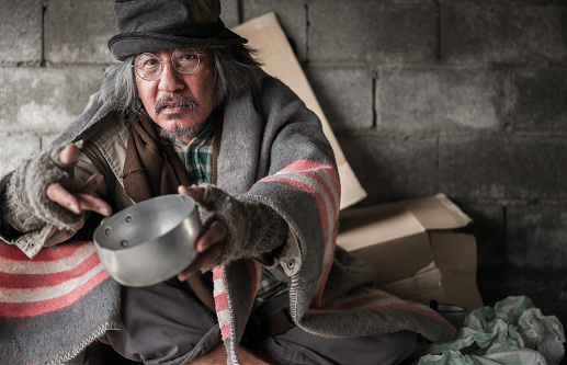 Old man beggar homeless is gray hair wears glasses and hat sitting on cardboard in shelter holding empty bowl for asks donate food and money on street. Senior poor male poverty hungry and sad concept.