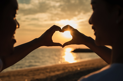 Silhouette of happy young couple forming a heart shape with their hands together on the beach while golden sunset time and sea background. Asian man and woman use hand heart shape for love at seaside.