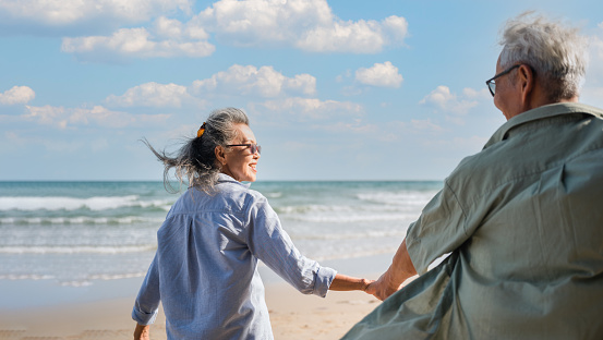Happy senior woman is enjoying and fun holding hand husband and pull to seaside on beach in vacation summer. Mature couple retired wear sunglasses hand in hand and relax walking for healthy at shore.