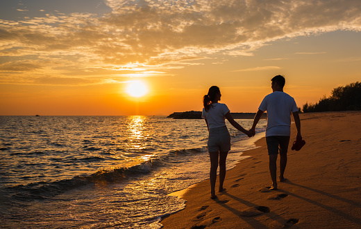 Silhouette of happy Asian couple hands holding and walking together on the beach while golden sunset time evening. Young in love romantic and relaxing on summer vacation in seaside. Honeymoon concept.