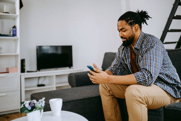 istock Happy Black Young Man Uses Smartphone while Sitting on a Sofa at Home. 1388252892