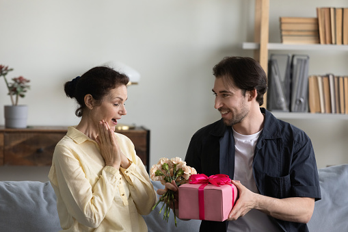 Happy excited mature mother getting present and flowers from grateful grown son, celebrating international women day, birthday, mothers day, receiving gift wrap, bouquet