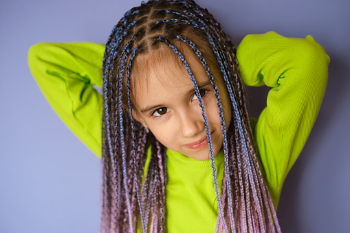 Nice sweet sweet girl with blue afro-pigtails dreadlocks. A child in a bright light green jacket on a Very Peri color background looks at the camera. Fashion and style
