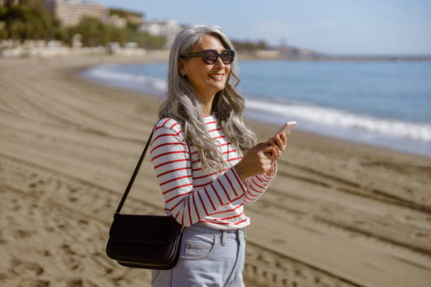 Elegant Asian lady standing outdoors by seafront Smiling mature woman in sunglasses with stylish handbag and mobile phone walking outdoors against backdrop of picturesque seascape older woman purse stock pictures, royalty-free photos & images