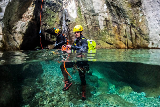 Two male canyoning adventurers seen in a beautiful canyon with crystal clear water while rappelling down the waterfall and one is holding the rope.