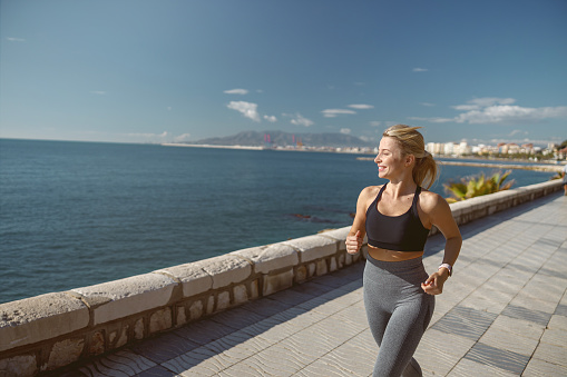 Caucasian good looking woman in sportswear keeping fit and healthy while running on sidewalk along seaside promenade