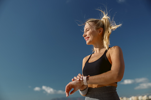 Side view of sporty blonde woman in training clothes adjusting fitness tracker during daily fitness training outdoors