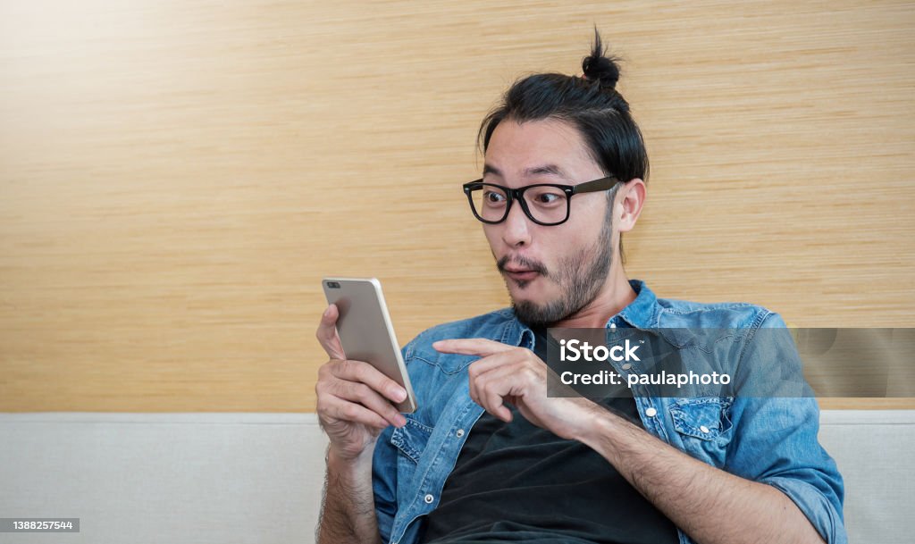 Portrait of happy young long hair nerd asian man wearing glasses typing sms on smart phone lifestyle in living room. Technology communication with friends family work, fun and surprise concept Nerd Stock Photo