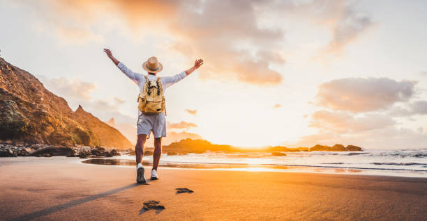 Happy man with hands up enjoying wellbeing and freedom at the beach - Male with backpack traveling in the nature with sunrise view - Healthy lifestyle, happiness and travel concept stock photo