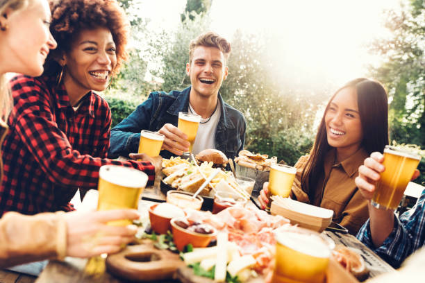 grupo de pessoas multiétnicas tendo jantar no quintal juntos - amigos millennials sentados à mesa de bar brindando copos de cerveja no jardim do pub da cervejaria - happy hour, intervalo de almoço e conceito de juventude - asian meal - fotografias e filmes do acervo