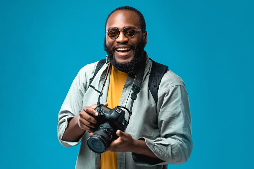 Portrait African-American man at the studio with colored background. Black person wearing sunglasses and blue t-shirt. Traveling and tourism concept. Photographer with professional digital camera