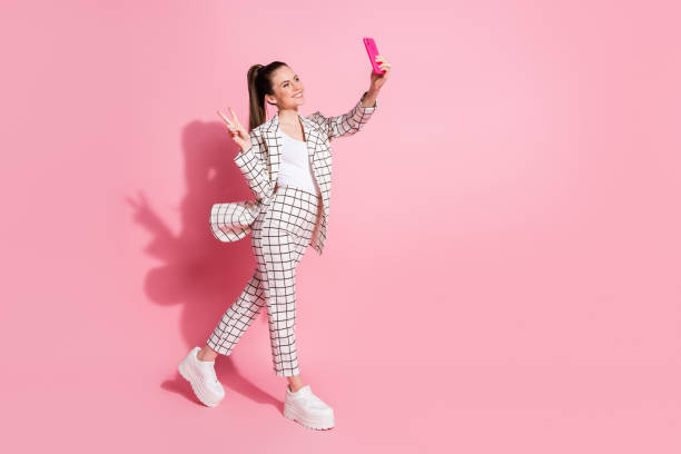 photo portrait full body view of woman taking selfies on the go showing v-sign isolated on pastel pink colored background - influencer stockfoto's en -beelden