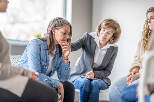 A female Therapist reaches out her hand and places it on the shoulder of a young woman to comfort her during a group therapy session.  The woman is dressed casually and appears distraught as she rests her chin in her hand.