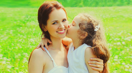 Portrait of little child daughter kissing her happy smiling mother on the grass in summer park