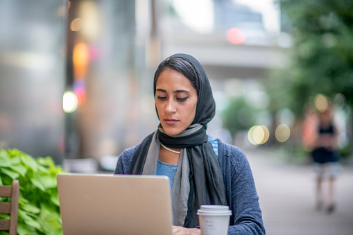 A young Muslim woman is seen sitting at an outdoor café table as she works away on her laptop.  She is dressed casually, has a Hijab on and a coffee beside her as she takes in the city life.