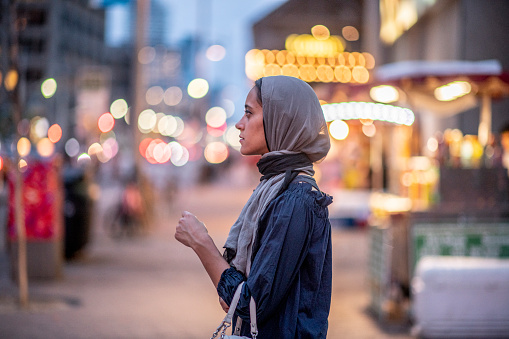 A young beautiful Muslim woman is seen walking the downtown streets at dusk, as she takes in the city life.  She is dressed casually, has a Hijab on and a purse on her arm as she walks the streets with a neutral expression on her face.