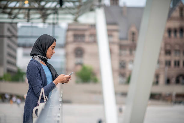 A young Muslim woman is seen walking the street as she makes her way into work in the morning.  She is dressed semi-casually, is wearing a Hijab, has a purse on her arm and her cell phone in hand as she checks her text messages.