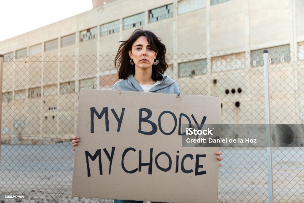 Young caucasian woman looking at camera with serious expression holding a cardboard sign: My body my choice Young caucasian woman looking at camera with serious expression holding a cardboard sign: My body my choice. Feminism activist concept. Protest Stock Photo