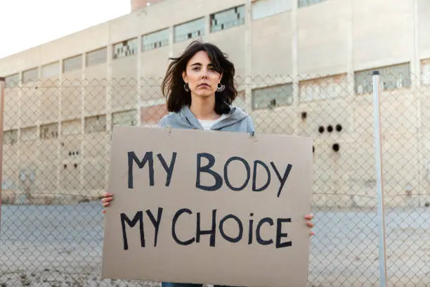 Photo of Young caucasian woman looking at camera with serious expression holding a cardboard sign: My body my choice