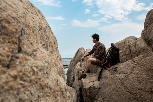 A young solo tourist is sitting on a rock overlooking the sea. Portrait.