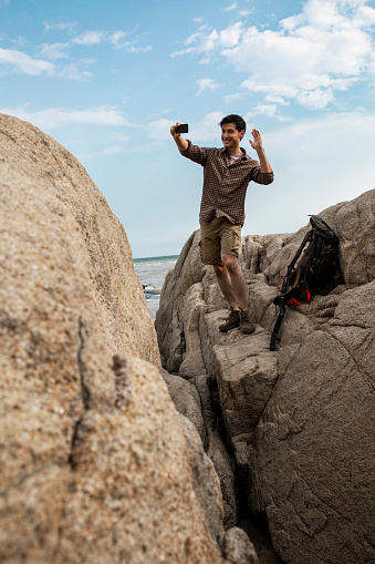 A young male outdoor traveler is taking a selfie while standing on a beach rock. Portrait.
