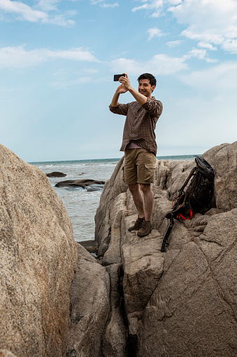 A young male outdoor traveler is standing on a beach rock and taking pictures of the scenery. Portrait.