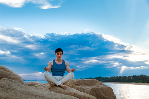Beneath the blue sea and sky clouds, a muscular man is taking the lotus position on a rock. portrait.