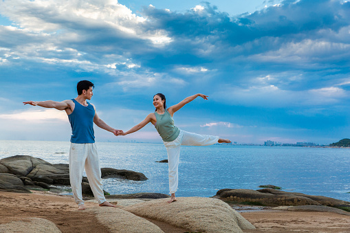 Beneath the spectacular sea and sky clouds, young couple is holding hands as they practice yoga stretches on an island rock. portrait.