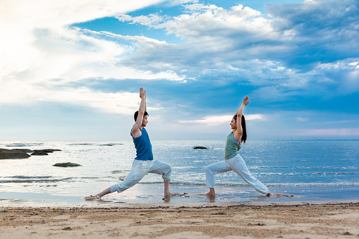 Under the blue clouds of sea and sky, young couple is holding hands as they practice yoga stretches on the beach. portrait.