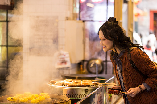 A young female college student selects fagao, a traditional Chinese snack, with a mobile phone and tanghulu at a snack street. In the southern of Chinese, Fagao, steamed rice cakes are famous.  Portrait.